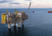 Equinor, Aker BP to Boost Norwegian Continental Shelf Output Offshore oil platform on three legs in the sea with a crane, ships visible in the distance.