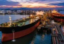 U.S. Oil Exports See Record Surge to Fill Global Supply Gap Oil tankers docked at a loading terminal at sunset, with pipelines and refinery structures in the background on calm water.
