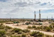 Devon Energy Tops Bid at Record $4B U.S. Oil and Gas Auction Oil drilling site in a desert with two tall rig towers, tanks, and interconnected pipelines in the foreground.