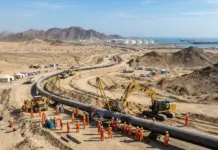 UAE Aims Second Oil Pipeline by 2027 to Bypass Hormuz Strait Construction crew in orange safety gear installing a large pipeline in a desert, with excavators, tents, and trucks nearby and a coastal port in the distance.