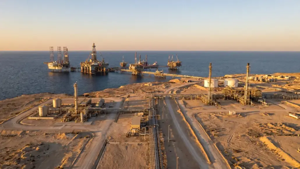 Aerial view of an offshore oil field with multiple platforms connected by pipelines along a desert coastline at sunset.