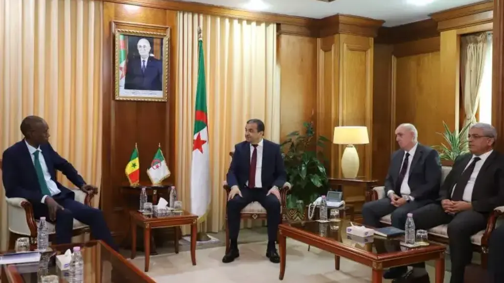 Four men in suits sit in a wood-paneled room for a formal diplomatic meeting, with flags and a portrait behind them.