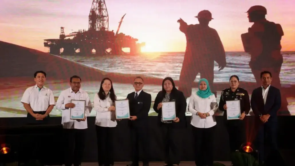 Eight people stand on a stage holding certificates during an award ceremony, with a sunset offshore oil rig backdrop behind them.