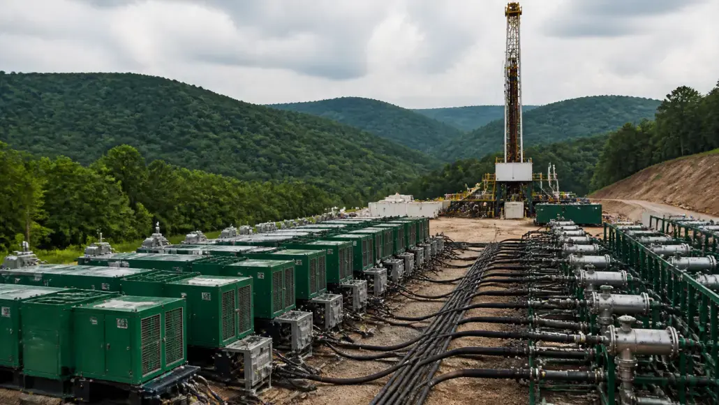 Row of green industrial generators with hoses leading to a tall oil-drilling rig in a hillside site with mountains in the background.